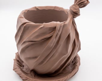 Puede incluir: Una maceta de cerámica marrón con forma de bolsa atada. La maceta tiene una abertura redonda y se asienta sobre una base de platillo a juego. La forma de bolsa presenta pliegues detallados y una parte superior anudada.