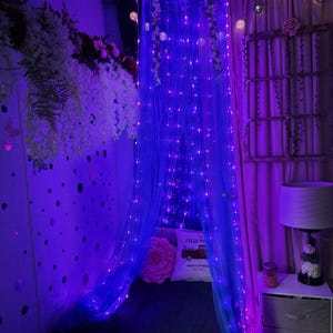Puede incluir: Una escena de dormitorio con una cama con dosel cubierta con una red azul e iluminada con luces de cadena. La cama está adornada con una almohada rosa y una almohada con un diseño de camión rojo. Arreglos florales y luces decorativas añaden ambiente.