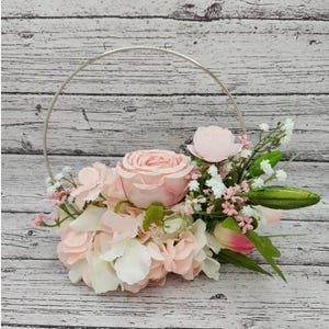 Puede incluir: Un arreglo floral rosa y blanco con una sola rosa rosa en el centro, rodeada de hortensias blancas y rosas y aliento de bebé. El arreglo se coloca en un aro de metal plateado.