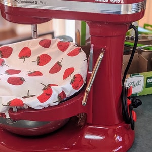 May include: A red stand mixer with a white bowl covered in a strawberry print fabric. The mixer has a silver handle and a black cord. The mixer is sitting on a gray countertop.