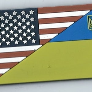 May include: A patch with the American flag and the Ukrainian flag, with the Ukrainian trident symbol in the upper right corner.