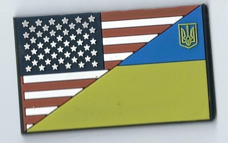 May include: A patch with the American flag and the Ukrainian flag, with the Ukrainian trident symbol in the upper right corner.