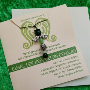 Peut inclure: Un pendentif en forme d'ange avec des perles vertes et des ailes argentées, présenté sur une carte avec du texte allemand. La carte présente un motif de cœur vert et les mots "ENGEL DER KRÖNENDEN ERFOLGE". Le collier est sur un fond vert.