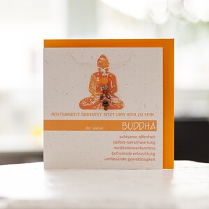 Può includere: Un biglietto di auguri quadrato con bordo arancione e centro color crema. La carta presenta un'illustrazione arancione di un Buddha seduto e testo in tedesco. La carta è su una superficie bianca.