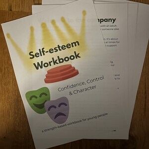 Può includere: Una pila di quaderni bianchi con il titolo "Self-esteem Workbook" in grandi lettere in grassetto. La copertina presenta una corona gialla, gradini rossi e maschere verdi e viola. È visibile anche il testo "Confidence, Control & Character".
