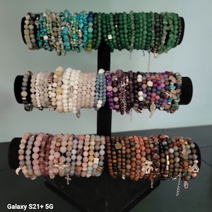 Puede incluir: Un expositor negro con una selección de pulseras de cuentas de colores. Las pulseras están hechas de varias piedras, incluyendo verde, blanco, morado y marrón. Las pulseras están apiladas en el expositor, creando una presentación visualmente atractiva.