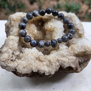 Könnte beinhalten: Ein dunkelgraues Perlenarmband mit einer Mischung aus matten und glänzenden Perlen. Das Armband liegt auf einer weißen und beigefarbenen Geode. Die Perlen sind rund und scheinen aus Stein zu sein.