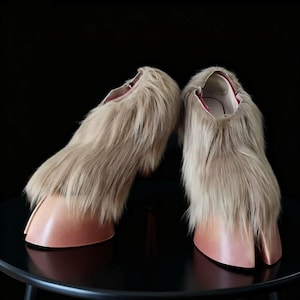May include: A pair of unique, avant-garde shoes. The shoes feature a light brown, furry upper and a pale pink, hoof-shaped toe. The interior lining is a deep red. The shoes are displayed on a black surface.