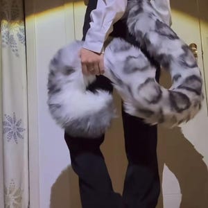Peut inclure: Une queue en fausse fourrure moelleuse dans les tons de gris et de blanc, tenue par une personne. La queue a une forme incurvée avec un motif tacheté, suggérant un accessoire de jeu ou de costume. La fourrure semble douce et volumineuse.