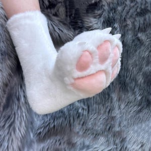 May include: White plush paw-shaped slipper with pink pads and white claws. The slipper is designed to resemble a cat's paw. The slipper is made of soft, fluffy material.