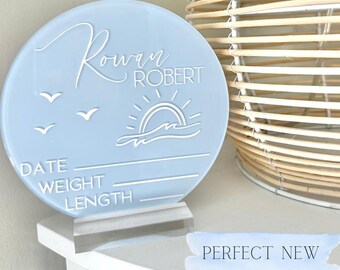 May include: A light blue, circular baby announcement sign with the name "Rowan Robert" in script. The sign features a sun and wave design, three birds, and spaces for date, weight, and length. It is displayed on a clear stand.