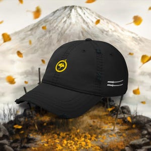 Puede incluir: Gorra de béisbol negra con acabado desgastado, con un emblema amarillo en la parte delantera y dos espadas katana blancas en el lateral. La gorra está colocada sobre un fondo de montaña y hojas cayendo.