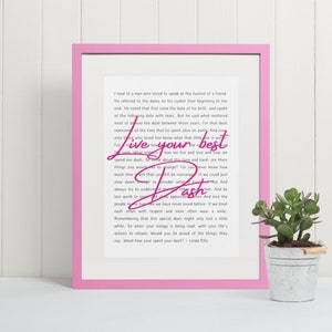 May include: A pink framed print with a white background and black text. The text reads "I read of a man who stood to speak at the funeral of a friend..." and then continues with a quote about living your best dash. The print is sitting on a white surface with a small potted plant in front of it.