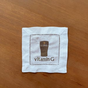 Może przedstawiać: Kwadratowa biała lniana podkładka z obszytą krawędzią. Środek przedstawia haftowany obrazek kufla Guinnessa z napisem "vitamin G" brązową nicią. Podkładka leży na drewnianej powierzchni.