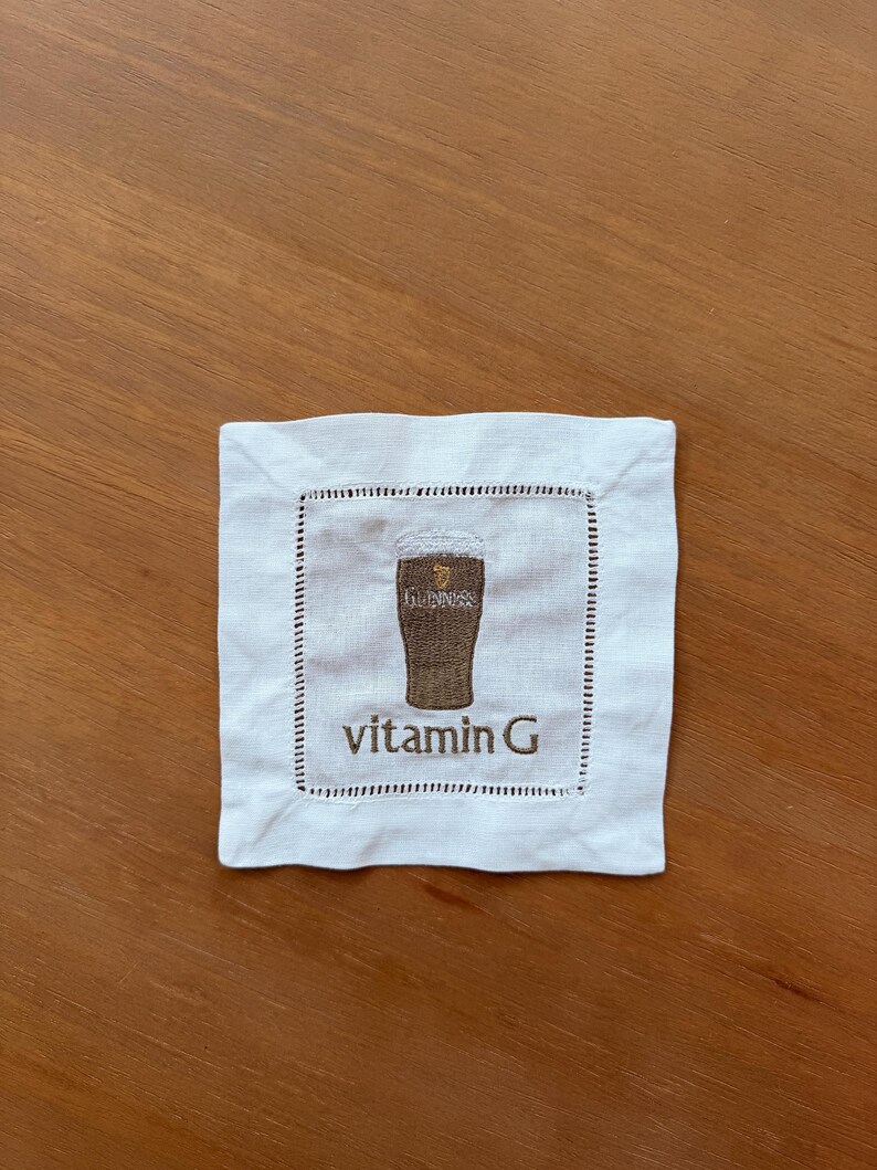 Może przedstawiać: Kwadratowa biała lniana podkładka z obszytą krawędzią. Środek przedstawia haftowany obrazek kufla Guinnessa z napisem "vitamin G" brązową nicią. Podkładka leży na drewnianej powierzchni.