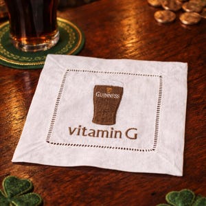 Może przedstawiać: Pintę piwa Guinness z białą pianą stoi na drewnianym barze obok białego lnianego podkładu. Podkładka ma haftowany kieliszek Guinnessa i napis "vitamin G". Obecne są r&oacute;wnież zielone dekoracje w kształcie koniczyny.