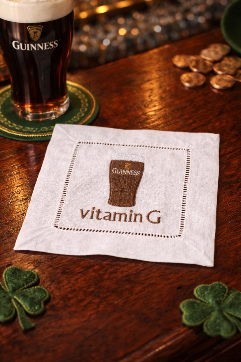 Może przedstawiać: Pintę piwa Guinness z białą pianą stoi na drewnianym barze obok białego lnianego podkładu. Podkładka ma haftowany kieliszek Guinnessa i napis "vitamin G". Obecne są r&oacute;wnież zielone dekoracje w kształcie koniczyny.