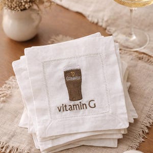 Może przedstawiać: Białe lniane serwetki koktajlowe z haftowanym wzorem szklanki Guinnessa i napisem "vitamin G". Serwetki są ułożone na beżowym lnianym obrusie. W tle kieliszek szampana i zapalona świeca.