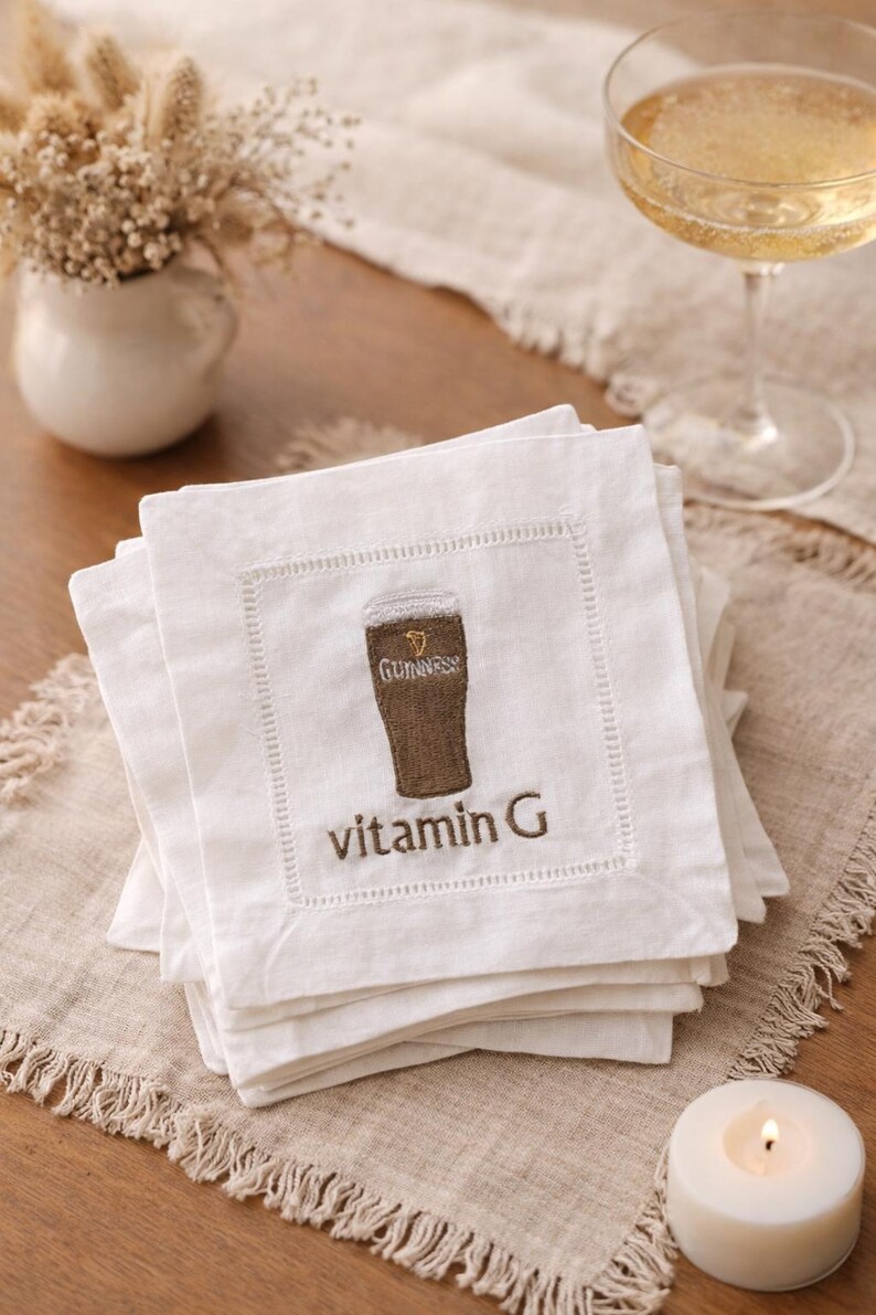 Może przedstawiać: Białe lniane serwetki koktajlowe z haftowanym wzorem szklanki Guinnessa i napisem "vitamin G". Serwetki są ułożone na beżowym lnianym obrusie. W tle kieliszek szampana i zapalona świeca.