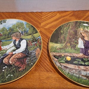 May include: Two decorative plates with gold rims. One plate depicts a man and a cat in a forest setting. The other plate shows a woman with a crown near a pond. Both plates have a fairytale theme.
