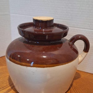 May include: A brown and white ceramic crock with a lid. The crock has a wide mouth and a handle on the side. The lid is brown with a white rim.