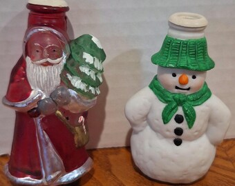 May include: Two ceramic Christmas figurines: Santa Claus and a snowman. The Santa figure is dressed in a red coat and holds a green Christmas tree. The snowman has a green hat and scarf, with black button details. Both are painted with bright colors.