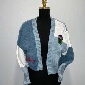 May include: A blue, white, and gray patchwork cardigan sweater. The sweater features a V-neck design and a cartoon character patch on the front. The word "Phillies" is embroidered in red on the front.