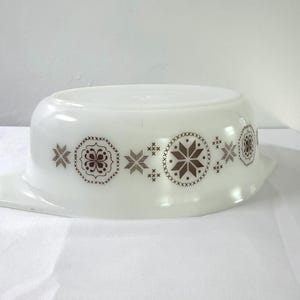 May include: A white oval milk glass casserole dish featuring a repeating brown pattern of stylized snowflakes and geometric cross-stitch motifs around the exterior. The dish has integrated side handles and a smooth, glossy finish throughout.