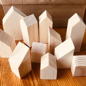 May include: A selection of small, unfinished wooden house-shaped blocks. The blocks have varying sizes and shapes, with angled roofs and rectangular bases. The natural wood grain is visible, arranged on a wooden surface.