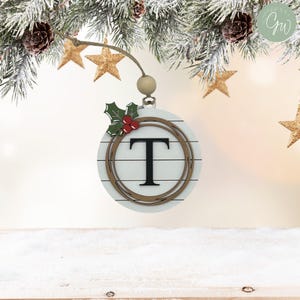 Puede incluir: Adorno navideño de madera blanca con la letra "T" negra en el centro, rodeada por un círculo marrón y adornada con hojas y bayas de acebo. Cuelga de una cuerda con una cuenta de madera. El fondo incluye ramas de pino, estrellas doradas y nieve.