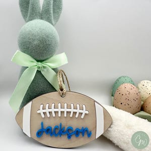 May include: Wooden football-shaped ornament with white stitching and the name "Jackson" in blue lettering. The ornament is hanging from a twine string.