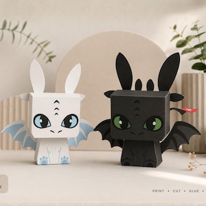 May include: Two papercraft dragon figures, one white with blue eyes and wings, and one black with green eyes and wings. The text CrazyPrint Paradise, PDF format, print, cut, glue, assemble, and level easy-intermediate are visible on the left side.