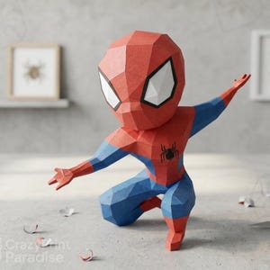 Puede incluir: Una figura de papel low-poly de un superhéroe en posición agachada. La figura presenta un diseño geométrico en rojo y azul con un emblema de araña negra en el pecho y grandes ojos blancos con bordes negros en la cabeza angular.