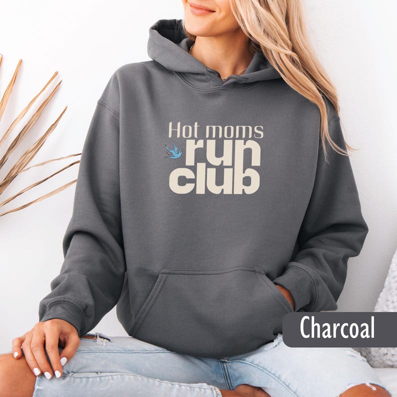 May include: Charcoal gray hoodie with the text "Hot moms run club" in a cream color. A small blue bird graphic is above the word "run". The hoodie has a front pocket and a drawstring hood.