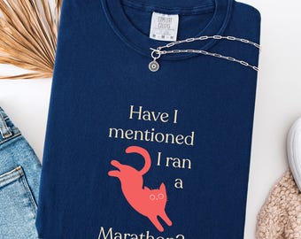 以下が含まれることがあります： 「Have I mentioned I ran a Marathon?」のテキストと赤い猫のグラフィックが特徴の、トゥルーネイビーブルーのスウェットシャツ。シルバーのネックレスがシャツの上にかけられています。スウェットシャツは白い表面に折りたたまれています。
