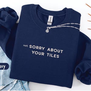 May include: A navy blue sweatshirt with the white text "not SORRY ABOUT YOUR TILES". A silver necklace with a small pendant is draped across the sweatshirt. The sweatshirt is folded on top of a pair of blue jeans.