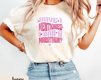 May include: Ivory-colored t-shirt with the text "HAVE I MENTIONED I RAN A MARATHON?" in purple and pink. The shirt has a relaxed fit and short sleeves. The text is in a playful, retro font.