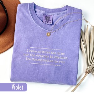May include: A violet-colored t-shirt with the text "I have neither the time nor the crayons to explain The Constitution to you." A gold necklace with a pendant is on the shirt. The shirt has a Comfort Colors tag.