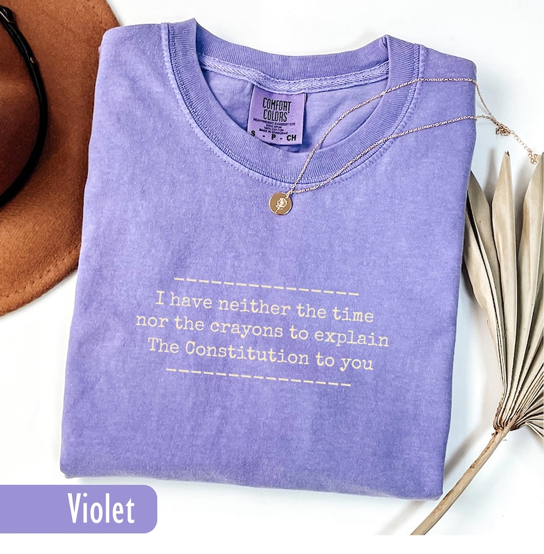 May include: A violet-colored t-shirt with the text "I have neither the time nor the crayons to explain The Constitution to you." A gold necklace with a pendant is on the shirt. The shirt has a Comfort Colors tag.