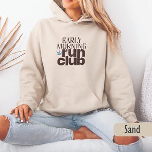 May include: A sand-colored hoodie with a front pocket and drawstring hood. The front features the text "EARLY MORNING run club" in a brown font with a blue bird graphic. The model is wearing light blue jeans.