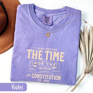 May include: A violet Comfort Colors t-shirt with the text "I HAVE NEITHER THE TIME NOR THE CRAYONS TO EXPLAIN The CONSTITUTION TO YOU." A gold necklace with a pendant rests on the shirt. A brown hat and dried plant are in the background.