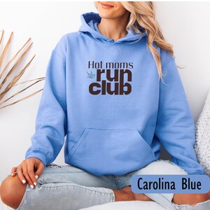 May include: Carolina Blue hoodie with the text "Hot moms run club" in brown. The hoodie has a front pocket and a hood. The text is accompanied by a small green bird graphic.