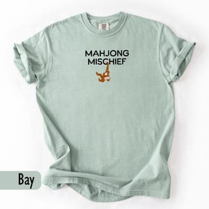 May include: A pale green t-shirt featuring the text "MAHJONG MISCHIEF" in black, with a cartoon orangutan holding a mahjong tile. The shirt has rolled-up sleeves and is a solid colour.