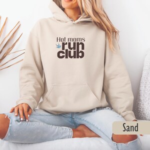 May include: A sand-colored hoodie with a drawstring hood and a front pocket. The hoodie features the text "Hot moms run club" in a dark brown font with a blue bird graphic. The model is wearing blue jeans.