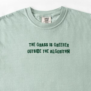 以下が含まれることがあります： ライトセージグリーンのクルーネックTシャツ。胸の中央には、ダークグリーンの掠れたフォントで「THE GRASS IS GREENER OUTSIDE THE ALGORITHM」というテキストがプリントされています。首元の内側にはComfort Colorsのブランドロゴラベルが付いています。