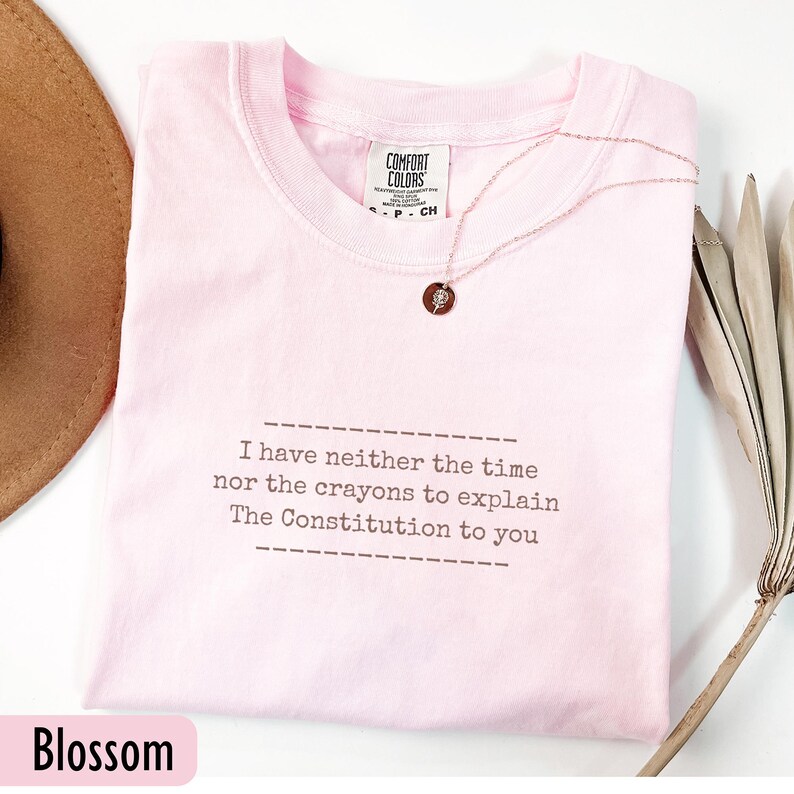 May include: A light pink t-shirt with the text "I have neither the time nor the crayons to explain The Constitution to you." A rose gold necklace with a pendant is on the shirt. The shirt is labeled "Blossom".