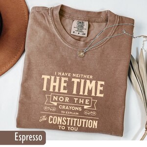 May include: Espresso-colored t-shirt with the text "I HAVE NEITHER THE TIME NOR THE CRAYONS TO EXPLAIN The CONSTITUTION TO YOU." A silver necklace with a small pendant rests on the shirt. The shirt is folded and has a Comfort Colors tag.