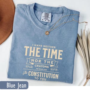 May include: A blue jean Comfort Colors t-shirt with the text "I HAVE NEITHER THE TIME NOR THE CRAYONS TO EXPLAIN The CONSTITUTION TO YOU." A gold necklace with a pendant rests on the shirt. The shirt is folded.