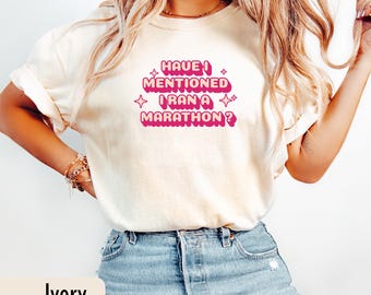 May include: Ivory-colored t-shirt with the text "HAVE I MENTIONED I RAN A MARATHON?" in pink. The text is in a retro font with star accents. The shirt is paired with blue jeans.