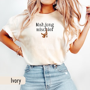 May include: Ivory-coloured t-shirt featuring the text "Mahjong mischief" in black, with a brown graphic below. The shirt is worn with blue jeans. The word "Ivory" is printed at the bottom of the image.